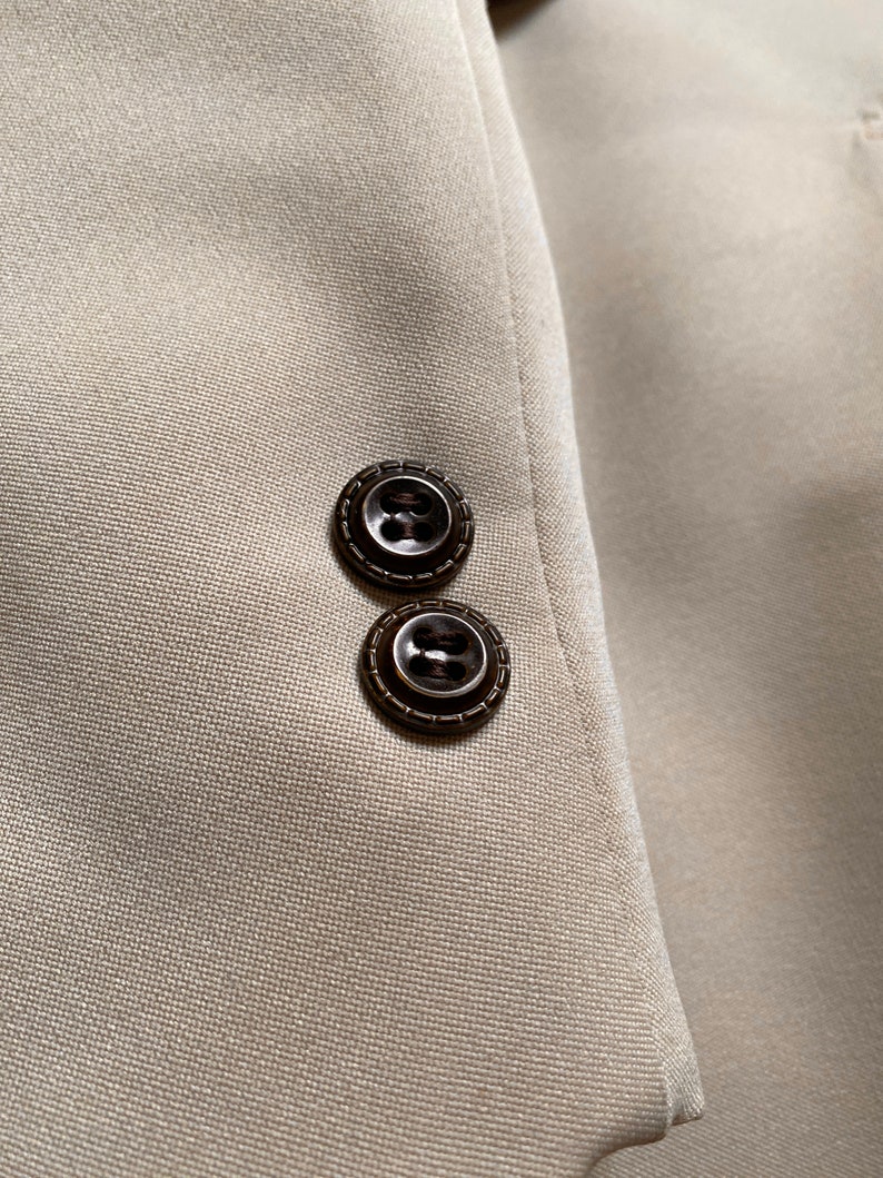 May include: Close-up of a beige blazer with two brown buttons on the sleeve.