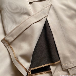 May include: A beige trench coat with a brown lining and a gold zipper. The coat has a belt and is made of a smooth fabric.