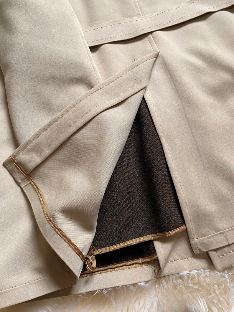 May include: A beige trench coat with a brown lining and a gold zipper. The coat has a belt and is made of a smooth fabric.