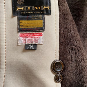 May include: A beige jacket with a brown fur lining. The jacket has a black label with the text "SIMS THE STORES FOR MEN TEXTURIZED POLYESTER" and a smaller label with the text "ADD 100% TEXTURIZED POLYESTER CARE INSTRUCTIONS: DRY CLEAN ONLY DO NOT STEAM OR PRESS PILE 42 REG."