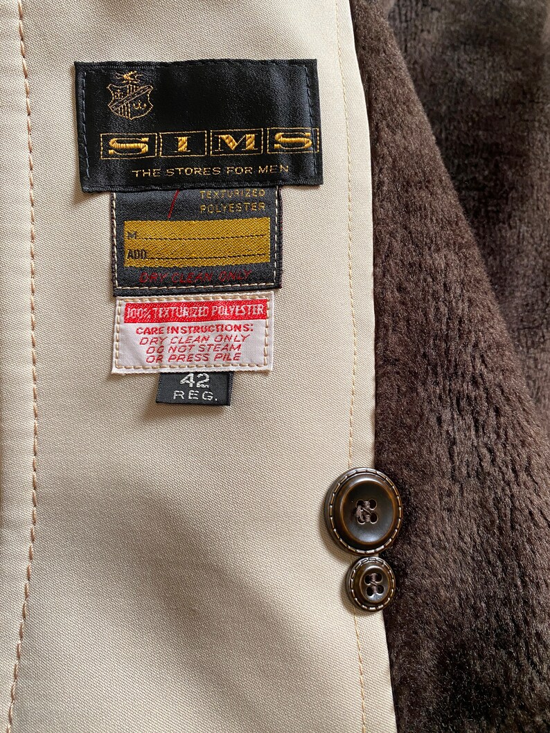 May include: A beige jacket with a brown fur lining. The jacket has a black label with the text "SIMS THE STORES FOR MEN TEXTURIZED POLYESTER" and a smaller label with the text "ADD 100% TEXTURIZED POLYESTER CARE INSTRUCTIONS: DRY CLEAN ONLY DO NOT STEAM OR PRESS PILE 42 REG."