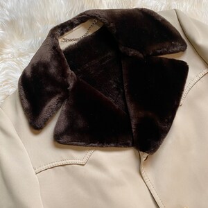 May include: A beige jacket with a brown faux fur collar. The jacket has a single button closure and a stitched detail on the front.