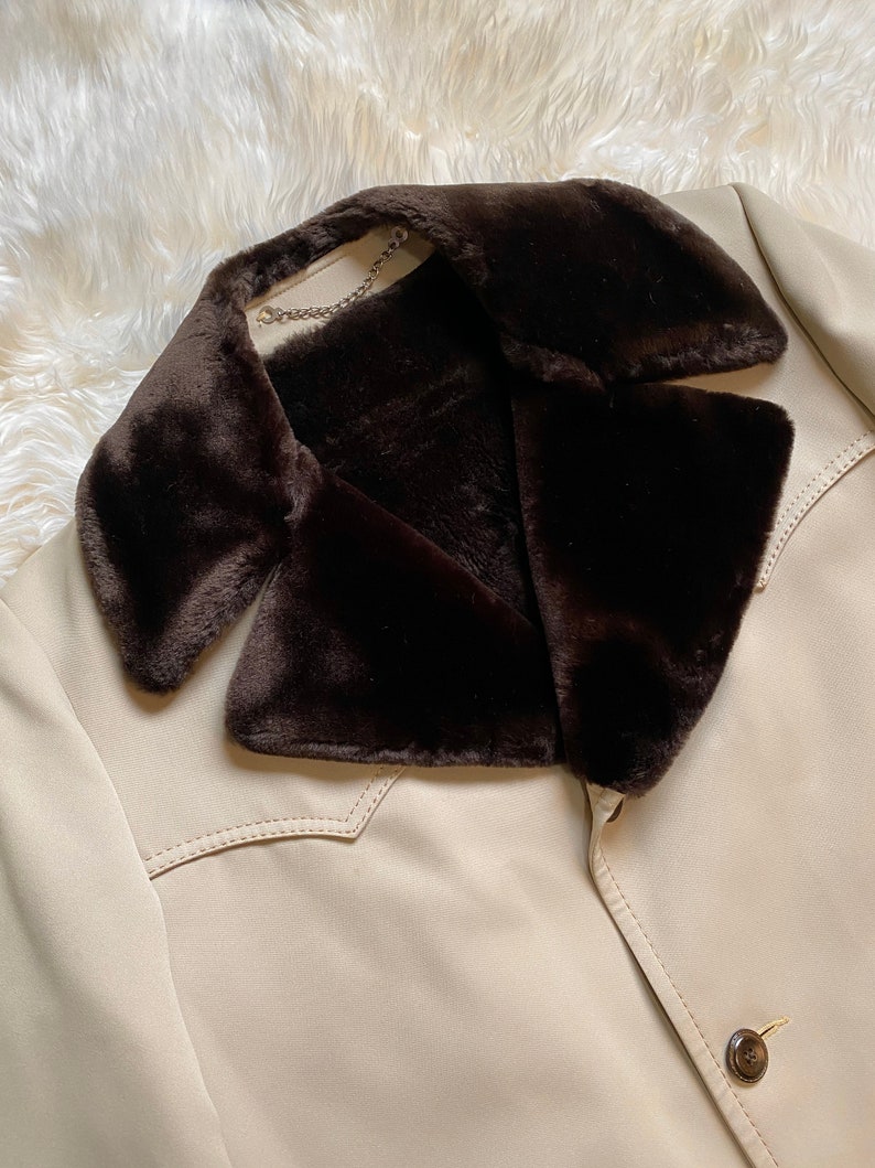 May include: A beige jacket with a brown faux fur collar. The jacket has a single button closure and a stitched detail on the front.