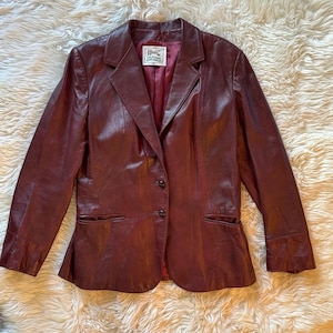 Puede incluir: Una chaqueta de cuero burdeos con un cierre de botón único y dos bolsillos. La chaqueta tiene una etiqueta que dice "H.I.C. California Leatherwear".