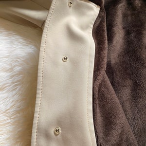 May include: Close-up of a beige jacket lining with brown faux fur. The lining has three silver buttons.