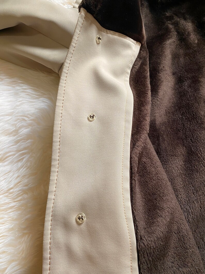 May include: Close-up of a beige jacket lining with brown faux fur. The lining has three silver buttons.