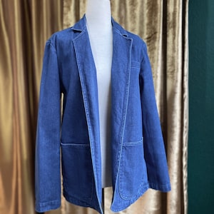 May include: A blue denim jacket with a classic blazer style. The jacket has a pointed collar, long sleeves, and two large pockets on the front.