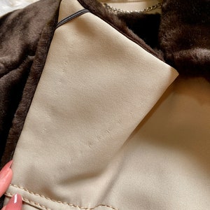 May include: Close-up of the inside of a brown faux fur jacket. The lining is a light beige color and has a black strap. The jacket has a zipper closure and a chain detail.