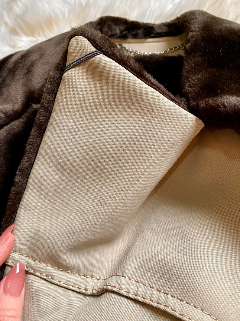 May include: Close-up of the inside of a brown faux fur jacket. The lining is a light beige color and has a black strap. The jacket has a zipper closure and a chain detail.