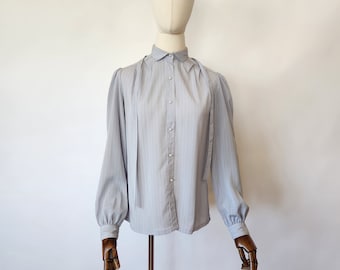 May include: A light gray long-sleeved blouse featuring a subtle vertical pinstripe pattern, a classic collar, and a front button placket with small white buttons. The garment includes long, full sleeves with buttoned cuffs and two long fabric ties.