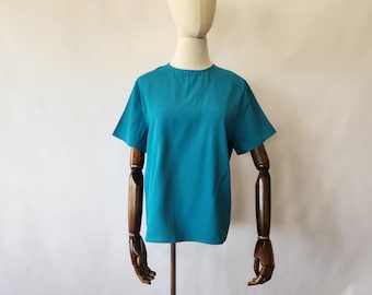 May include: A teal-colored short-sleeve top displayed on a mannequin. The top has a simple, round neckline and a loose fit. The mannequin has a neutral-colored head and wooden arms. The background is a plain white.