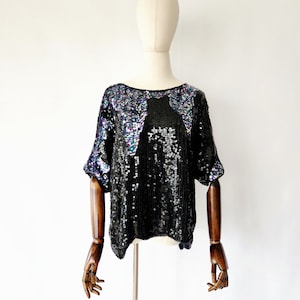 Pode incluir: Um top preto de lantejoulas com lantejoulas iridescentes. O top tem um decote redondo e mangas curtas.