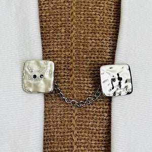 May include: Silver tone metal square shaped shawl clips with a chain connecting them. The clips have a hammered texture.
