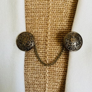 Pode incluir: Dois botões de metal ornamentados em tom dourado, conectados por uma corrente, contra um fundo de tecido de malha bege.