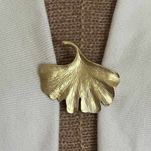 Puede incluir: Broche de metal dorado con forma de hoja de ginkgo. La broche se coloca sobre una tela blanca y un fondo de tela marrón texturizada. El diseño de la hoja tiene vetas detalladas.