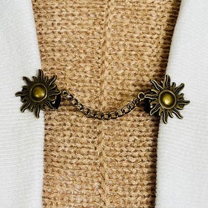 May include: A decorative bronze-toned sunburst brooch with a chain connecting two sun-shaped clasps. The brooch is set against a textured beige background and white fabric.