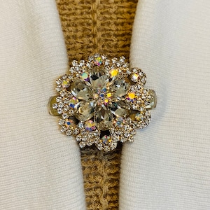 Op de afbeelding: Een goudkleurige servetring met een bloemmotief. De ring is bezet met heldere en iriserende strass-steentjes, wat een sprankelend effect creëert. De servetring houdt een witte stoffen servet vast.