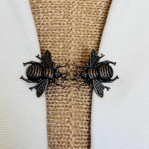 May include: A bronze bee-shaped clasp for fabric. The clasp has two detailed bee figures with wings and body segments, joined by a small chain. The background shows white fabric and a textured beige material.
