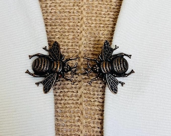 May include: A bronze-colored bee-shaped clasp, designed to fasten fabric. The clasp features two detailed bee figures with wings and body segments, connected by a small chain. The background shows white fabric and a textured beige material.