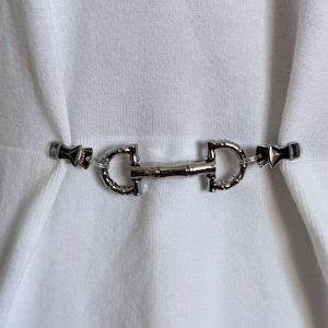 May include: A silver-coloured metal belt buckle with a bamboo-style centre and black accents. The buckle is attached to a white fabric, creating a contrast between the shiny metal and the soft texture of the material.