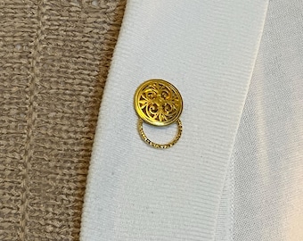 May include: Close-up of a pale blue cardigan featuring a gold-coloured button. The button is round with an ornate design and a small ring attached. The cardigan's fabric has a ribbed texture.
