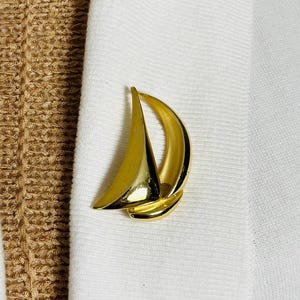 May include: A gold-toned sailboat brooch pin. The pin is a simple design with a single sail.