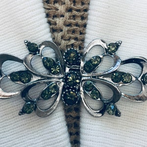 Op de afbeelding: Een zilveren broche in de vorm van een vlinder met groene strass steentjes. De broche is versierd en gedetailleerd, met een delicaat ontwerp.