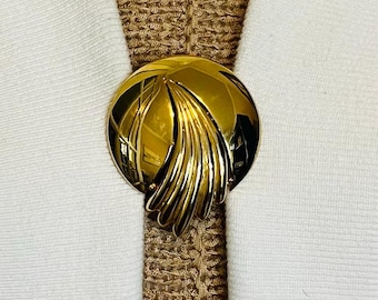 May include: A gold-colored, round brooch with a stylized leaf design. The brooch is set against a textured, tan fabric and white fabric background, creating a striking contrast. The brooch is a vintage accessory.