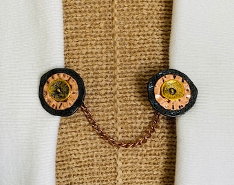 May include: A black and copper chain with two round, black and copper, decorative clasps. The clasps have a raised, gold-coloured centre.