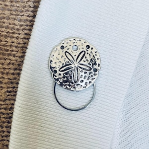 Puede incluir: Broche plateado con forma de dólar de arena con un anillo circular. El broche presenta un diseño de estrella de mar y pequeños detalles negros. Se fija a una tela de punto blanca.