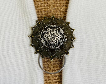 May include: A bronze-coloured metal clasp with a decorative design featuring a silver-coloured snowflake. The clasp has a ring on the bottom for attaching to fabric.