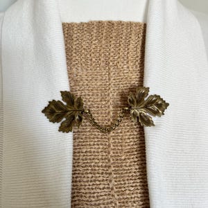 Puede incluir: Un broche de cardigan en forma de hoja de color dorado con una cadena que conecta las dos hojas. El broche está en un suéter marrón.