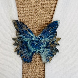 May include: A blue butterfly-shaped decorative item with gold accents. The butterfly has detailed wing patterns and is set against a white and tan background. The item appears to be a decorative accessory.