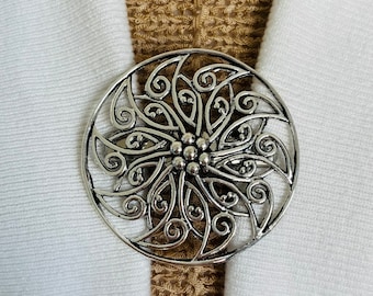 May include: A silver circular brooch with a decorative floral design. The brooch has a filigree pattern with swirls and a central flower.
