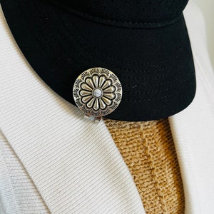 May include: A silver-colored, flower-shaped hat clip is attached to a black visor. The clip has a central, light-colored stone and a detailed, embossed design. The visor has a white Nike logo.
