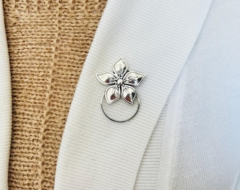 May include: A silver flower brooch with five petals and a central detail, pinned to a white cardigan. The brooch has a circular pin backing. The cardigan is worn over a beige knit sweater.