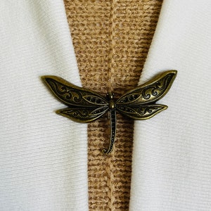 May include: A gold-toned dragonfly brooch with intricate details. The brooch is attached to a white and brown fabric background.