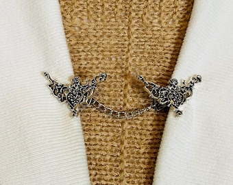 Peut inclure: Clip de pull en métal argenté avec un motif de papillon orné. Le clip est relié par une chaîne délicate et est présenté sur un tissu blanc sur un fond beige texturé. Le clip est conçu pour attacher le devant d'un pull ou d'un cardigan.
