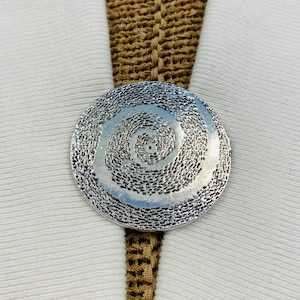 May include: A silver-colored, circular pendant with a textured spiral design. The pendant is set against a white fabric background with a brown, woven strap.