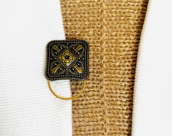 May include: A gold-toned metal clasp with an intricate design. The clasp is square-shaped and has a decorative pattern with a central flower motif.