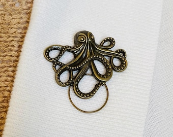 May include: A bronze octopus napkin ring. The octopus design features detailed tentacles and a circular ring. The napkin ring is displayed on a white cloth.