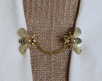 May include: A gold-toned metal cardigan clip featuring two bee-shaped fasteners connected by a delicate chain. Each bee is embellished with clear crystals on the wings, black stones on the abdomen, and a smooth, round, light brown faux pearl head.