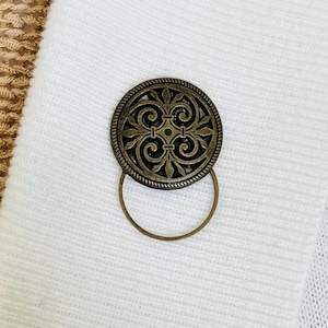 May include: A round, antique brass button with an ornate, swirling design and a small loop. The button is set against a white fabric background, highlighting its intricate details and aged appearance.