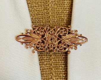 May include: A rose gold-colored metal brooch with an ornate, symmetrical design. The brooch features a central floral element and intricate scrollwork. It is displayed on a white fabric and a textured gold fabric.