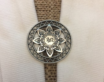 May include: A silver circular brooch with a detailed floral design and a central star with the letter "S" inside. The brooch is attached to a brown fabric strap.