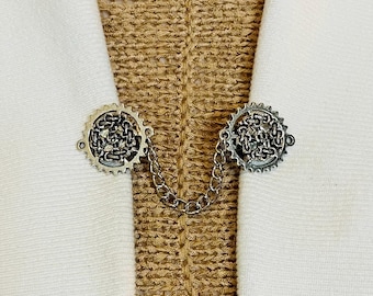 Peut inclure: Deux engrenages métalliques argentés reliés par une chaîne, portés comme une broche décorative sur un tissu en maille marron.