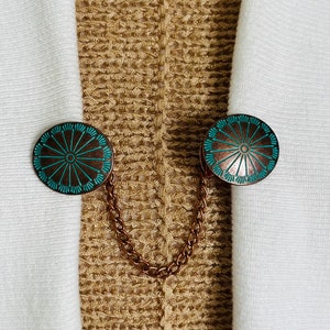 May include: A pair of copper-toned metal brooches with a turquoise blue design. The brooches are connected by a chain.
