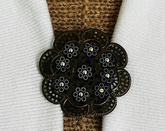 May include: A large, ornate, circular, bronze-colored metal brooch with a floral design. The brooch is decorated with small, black, round stones.