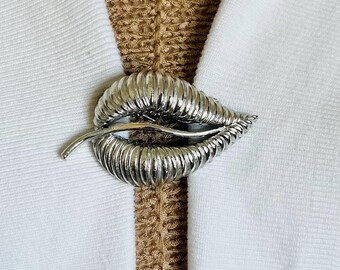 May include: A silver-toned leaf brooch with a textured, ribbed design. The brooch is shaped like a leaf with a curved stem and is set against a white fabric background. The brooch is a fashion accessory.