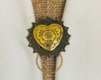 May include: A gold heart-shaped brooch with intricate details and a decorative ring attached. The brooch is on a brown fabric background.